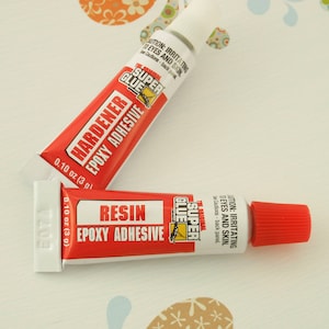 Puede incluir: Dos tubos de adhesivo epoxi Super Glue. El tubo de la izquierda est&aacute; etiquetado como "Hardener Epoxy Adhesive" y el tubo de la derecha est&aacute; etiquetado como "Resin Epoxy Adhesive". Ambos tubos son rojos y blancos con texto negro y tienen 0,10 oz (3 g).