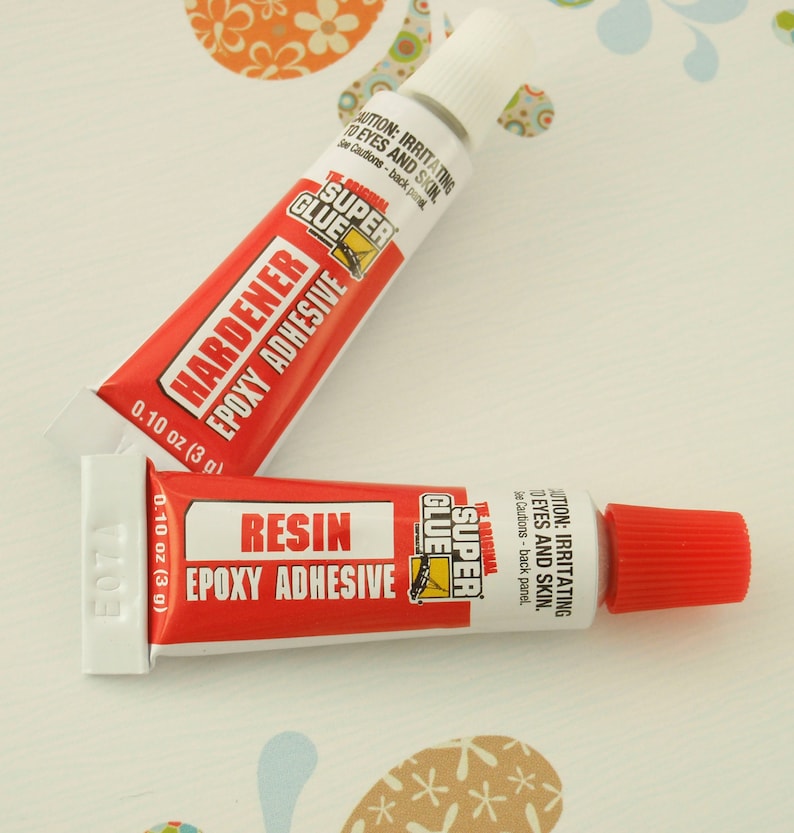 Puede incluir: Dos tubos de adhesivo epoxi Super Glue. El tubo de la izquierda est&aacute; etiquetado como "Hardener Epoxy Adhesive" y el tubo de la derecha est&aacute; etiquetado como "Resin Epoxy Adhesive". Ambos tubos son rojos y blancos con texto negro y tienen 0,10 oz (3 g).