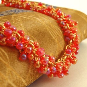 May include: A necklace featuring a dense cluster of small, translucent red beads with an iridescent finish. The beads are intricately woven onto a gold-toned metal chain, creating a textured, multi-layered appearance that catches the light.