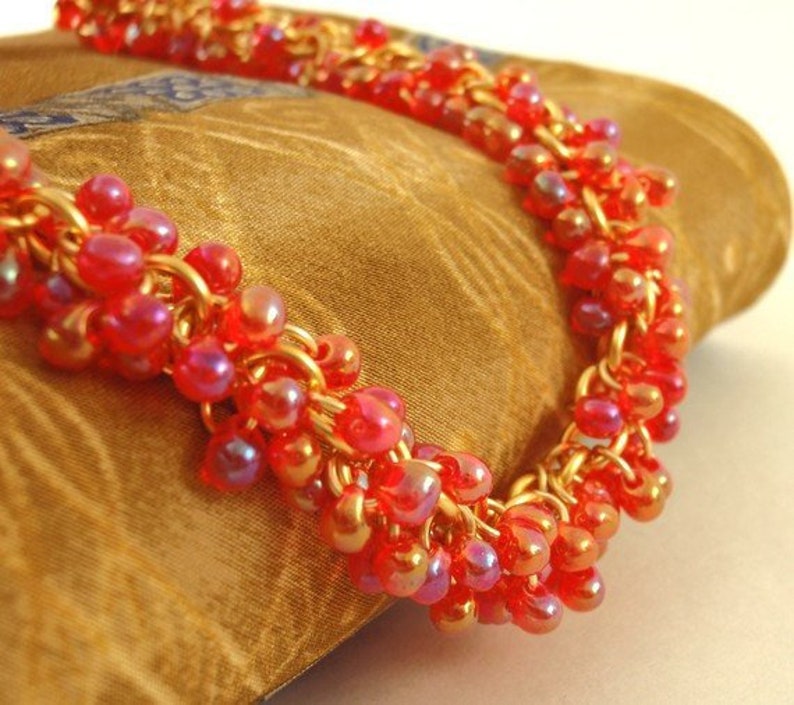May include: A necklace featuring a dense cluster of small, translucent red beads with an iridescent finish. The beads are intricately woven onto a gold-toned metal chain, creating a textured, multi-layered appearance that catches the light.