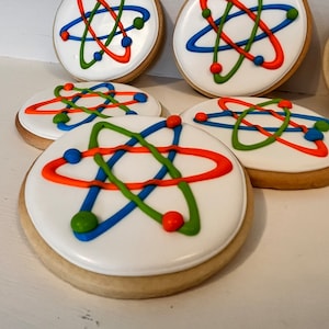 May include: Round sugar cookies with white icing, decorated with colorful atom designs in orange, blue, and green. The cookies have a light brown edge and are arranged on a white surface. The atom design features curved lines and small ball accents.