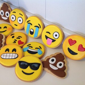 May include: A collection of emoji-themed cookies, including smiling faces, crying faces, and a winking face with a tongue sticking out. The cookies are yellow with various facial expressions and are decorated with icing. There is also a cookie shaped like a poop emoji.