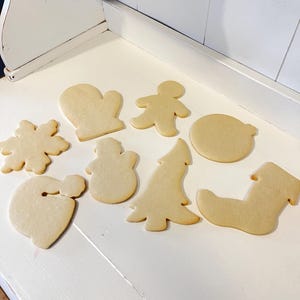 Puede incluir: Una colecci&oacute;n de galletas de az&uacute;car sin decorar en varias formas navide&ntilde;as, que incluyen un copo de nieve, un guante, un hombre de jengibre, un mu&ntilde;eco de nieve, un &aacute;rbol de Navidad, un gorro de Pap&aacute; Noel, un c&iacute;rculo y un calcet&iacute;n. Las galletas son de color marr&oacute;n claro.