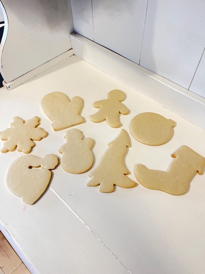 Puede incluir: Una colecci&oacute;n de galletas de az&uacute;car sin decorar en varias formas navide&ntilde;as, que incluyen un copo de nieve, un guante, un hombre de jengibre, un mu&ntilde;eco de nieve, un &aacute;rbol de Navidad, un gorro de Pap&aacute; Noel, un c&iacute;rculo y un calcet&iacute;n. Las galletas son de color marr&oacute;n claro.