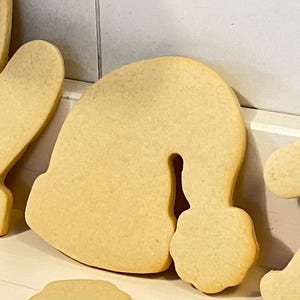 Puede incluir: Primer plano de varias galletas de az&uacute;car de color marr&oacute;n claro sin adornos. Una galleta tiene forma de gorro de Pap&aacute; Noel, con la parte superior redondeada y un pomp&oacute;n. Las otras galletas son parcialmente visibles. Las galletas est&aacute;n sobre una superficie blanca.