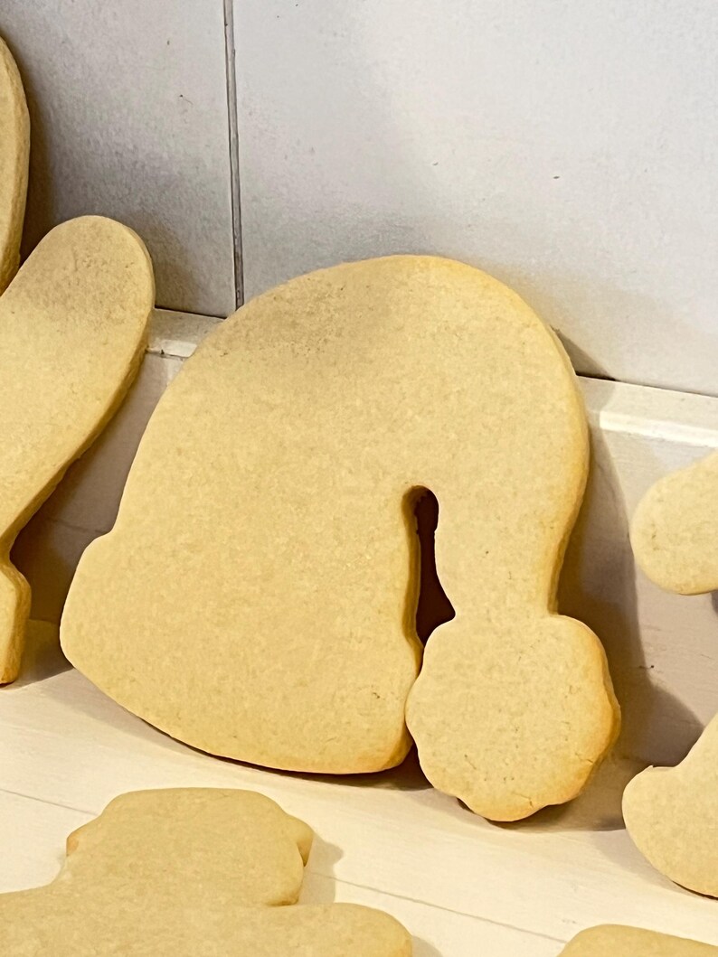 Puede incluir: Primer plano de varias galletas de az&uacute;car de color marr&oacute;n claro sin adornos. Una galleta tiene forma de gorro de Pap&aacute; Noel, con la parte superior redondeada y un pomp&oacute;n. Las otras galletas son parcialmente visibles. Las galletas est&aacute;n sobre una superficie blanca.