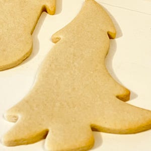 Puede incluir: Primer plano de varias galletas de az&uacute;car reci&eacute;n horneadas, de color marr&oacute;n claro. Una galleta tiene forma de &aacute;rbol de Navidad, con un contorno detallado. Las galletas est&aacute;n sobre una superficie blanca, lo que sugiere un tema festivo.