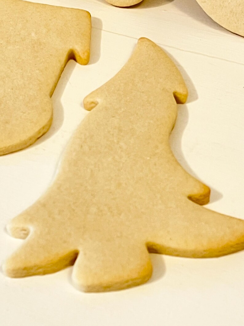 Puede incluir: Primer plano de varias galletas de az&uacute;car reci&eacute;n horneadas, de color marr&oacute;n claro. Una galleta tiene forma de &aacute;rbol de Navidad, con un contorno detallado. Las galletas est&aacute;n sobre una superficie blanca, lo que sugiere un tema festivo.