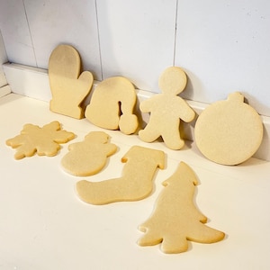 Puede incluir: Un surtido de galletas navide&ntilde;as sin decorar, de color marr&oacute;n claro, en varias formas, incluyendo un guante, gorro de Pap&aacute; Noel, hombre de jengibre, adorno, copo de nieve, mu&ntilde;eco de nieve, calcet&iacute;n y &aacute;rbol de Navidad. Las galletas est&aacute;n dispuestas sobre una superficie blanca.