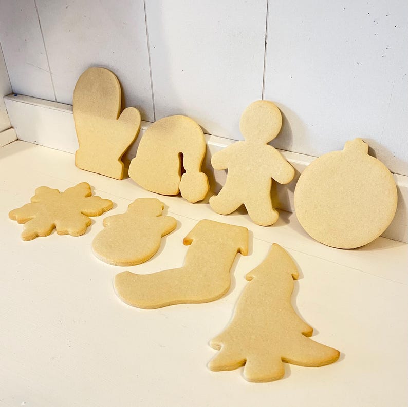 Puede incluir: Un surtido de galletas navide&ntilde;as sin decorar, de color marr&oacute;n claro, en varias formas, incluyendo un guante, gorro de Pap&aacute; Noel, hombre de jengibre, adorno, copo de nieve, mu&ntilde;eco de nieve, calcet&iacute;n y &aacute;rbol de Navidad. Las galletas est&aacute;n dispuestas sobre una superficie blanca.