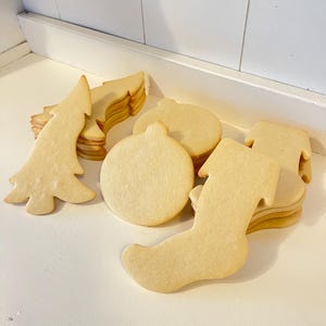 Puede incluir: Una colecci&oacute;n de galletas de az&uacute;car sin hornear con tem&aacute;tica navide&ntilde;a. Las formas incluyen un &aacute;rbol de Navidad, una bola y un calcet&iacute;n. Las galletas son de color marr&oacute;n claro y est&aacute;n apiladas en peque&ntilde;os montones sobre una superficie blanca.