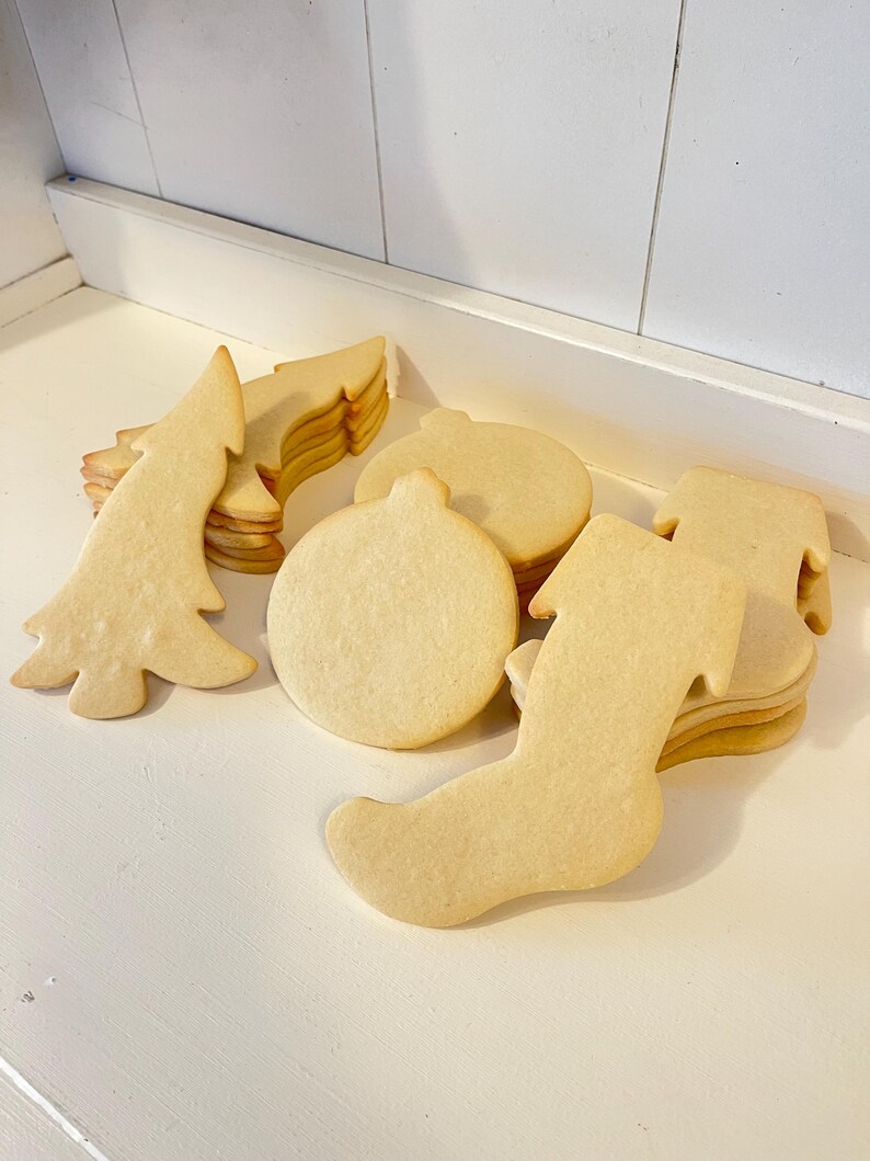 Puede incluir: Una colecci&oacute;n de galletas de az&uacute;car sin hornear con tem&aacute;tica navide&ntilde;a. Las formas incluyen un &aacute;rbol de Navidad, una bola y un calcet&iacute;n. Las galletas son de color marr&oacute;n claro y est&aacute;n apiladas en peque&ntilde;os montones sobre una superficie blanca.
