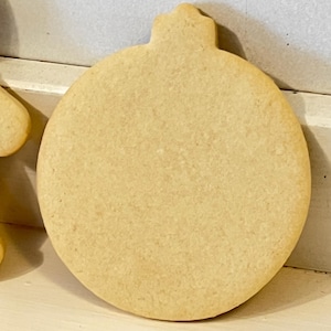Puede incluir: Una galleta grande, sin decorar, con forma de adorno navide&ntilde;o. La galleta es de color marr&oacute;n claro y tiene una forma redondeada con una peque&ntilde;a parte superior decorativa. La galleta es probablemente casera y lista para decorar.