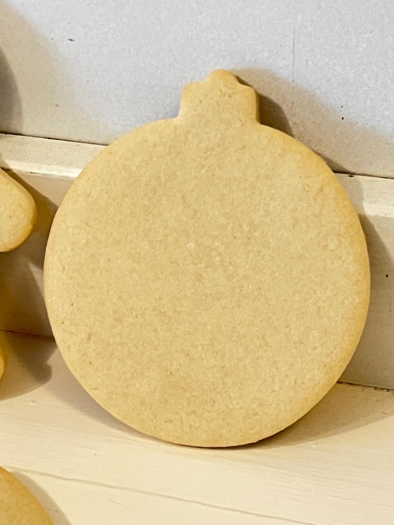 Puede incluir: Una galleta grande, sin decorar, con forma de adorno navide&ntilde;o. La galleta es de color marr&oacute;n claro y tiene una forma redondeada con una peque&ntilde;a parte superior decorativa. La galleta es probablemente casera y lista para decorar.
