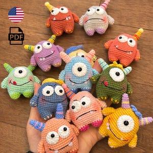 Może przedstawiać: Kolekcja kolorowych, szydełkowych zabawek-potworów w różnych kolorach, w tym pomarańczowym, fioletowym, niebieskim i zielonym. Każdy potwór ma unikalny design z rogami, oczami i uśmiechniętą twarzą. Zabawki są ręcznie robione.