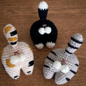 Op de afbeelding: Drie gehaakte kattenkontornamenten. Eén is zwart met een witgepunte staart, een andere is lichtgrijs met zwarte strepen, en de derde is bruin met zwarte en gouden accenten. Elk heeft een klein roze kruis en twee witte pompons.