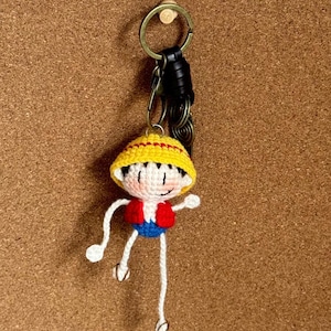 May include: Crocheted keyring featuring a cartoon character with a yellow hat, red vest, and blue shorts. The keyring has a metal ring and a black leather strap. The character is white, with black eyes and a smiling expression.