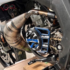 Può includere: Primo piano di un motore di moto con un tubo di scarico argentato e una copertura protettiva nera e blu. La copertura presenta il testo "DERBI" e "Stage6" in bianco e blu. È visibile una catena dorata.