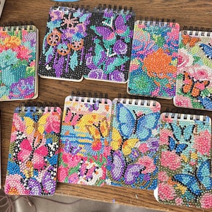 May include: A collection of small, spiral-bound notebooks with colorful, beaded designs. The covers feature various floral and butterfly patterns in shades of pink, blue, purple, and orange. The notebooks are arranged on a wooden surface.