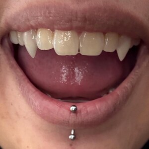 May include: Close-up of a mouth with vampire fangs. The upper teeth are sharp and pointed, with a tongue visible inside the mouth. A lip piercing is present on the lower lip.