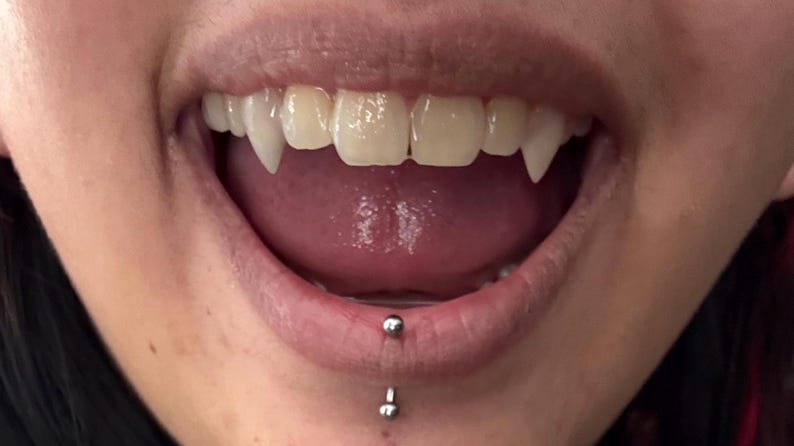 May include: Close-up of a mouth with vampire fangs. The upper teeth are sharp and pointed, with a tongue visible inside the mouth. A lip piercing is present on the lower lip.