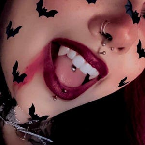 May include: Close-up of a person's mouth with dark red lipstick and fangs. The person has multiple facial piercings and black bat stickers around the face. The tongue has a silver barbell piercing.
