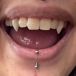 May include: Close-up of a mouth with sharp, pointed vampire fangs. The teeth are white, contrasting with the pink tongue and inner mouth. A lip piercing is visible below the lower lip.