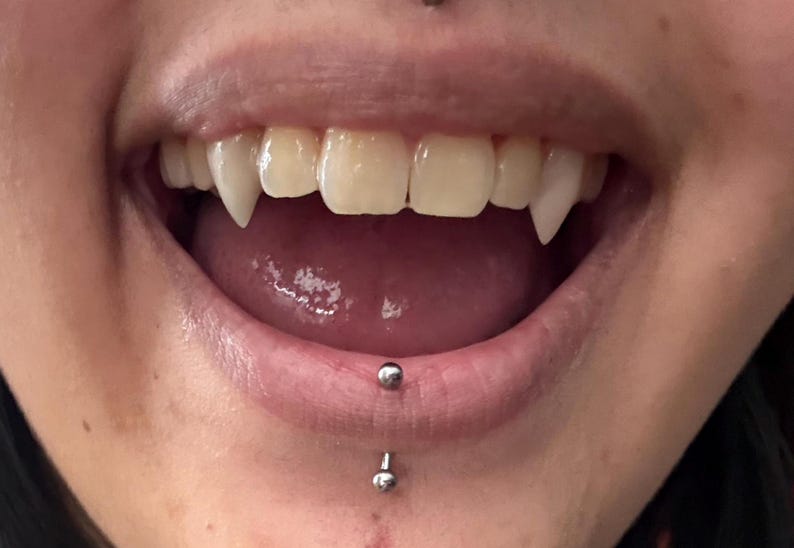May include: Close-up of a mouth with sharp, pointed vampire fangs. The teeth are white, contrasting with the pink tongue and inner mouth. A lip piercing is visible below the lower lip.