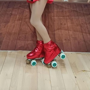 Puede incluir: Un par de patines de ruedas rojos con ruedas blancas y detalles verdes. Los patines tienen un acabado met&aacute;lico y los lleva puesta una persona de pie sobre un suelo de madera.