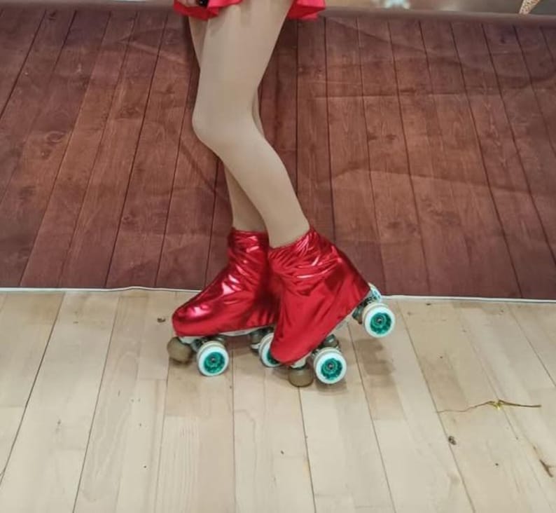 Puede incluir: Un par de patines de ruedas rojos con ruedas blancas y detalles verdes. Los patines tienen un acabado met&aacute;lico y los lleva puesta una persona de pie sobre un suelo de madera.