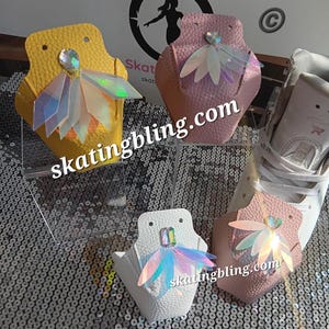 May include: A display of skating bling accessories in yellow, pink, and white. Each accessory features a decorative iridescent wing design and a gemstone. The website address skatingbling.com is visible on the accessories.