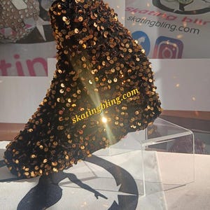 Könnte beinhalten: Ein schwarzer Rollschuhstiefel, der mit goldenen Pailletten bedeckt ist. Der Stiefel wird auf einem transparenten Acrylständer präsentiert. Der Text "skatingbling.com" ist auf dem Stiefel zu sehen. Der Hintergrund zeigt Bilder von anderen Schlittschuhen.
