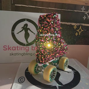 May include: A pair of roller skates with a black and pink sequined boot cover. The boot cover has a rainbow iridescent sheen. The skates are on a white surface with a black and white logo that says "skating bling.com".