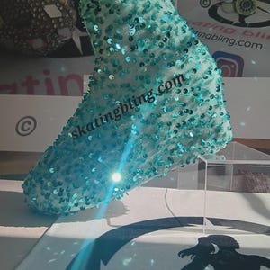 May include: A turquoise blue ice skate boot cover with sequins. The cover has the text "skatingbling.com" on it.