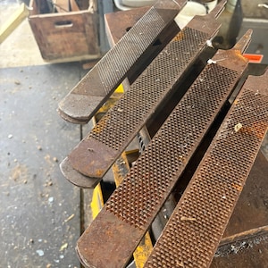 May include: Four rusty metal files with textured surfaces are arranged diagonally. The files have a rectangular shape with a handle-like extension at the top. The tools are likely used for shaping or smoothing materials. The background shows workshop elements.