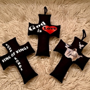 May include: Three black fabric cross-shaped pillows with different designs. One cross has the text "God is Love" with a red heart. Another cross has the text "Lord King of Kings Lord". The third cross has a picture of Jesus with doves flying around him.
