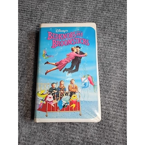 May include: A white plastic VHS case for Disney's Bedknobs and Broomsticks. The cover features a blue background with pink text, a flying couple, three children sitting on a brass bed frame, and several colorful cartoon fish swimming nearby.