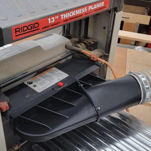 May include: A close-up of a Ridgid 13" Thickness Planer with a black dust collection hose attached to the dust port. The planer has a large capacity and high performance.