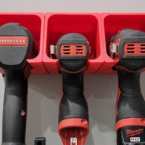 May include: A red 3-slot drill holder with three black and red power drills. The drills are labeled "BRUSHLESS" and "Milwaukee M12". The holder is mounted on a light gray wall.