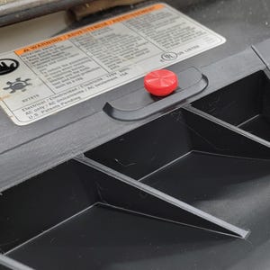 May include: Close-up of a black plastic surface with a red button. The surface has several compartments and a warning label with the text "A WARNING / ADVERTENCIA / AVERTISSEMENT" and "Electrical / Electricidad / AC seulement 120V 60Hz USA".