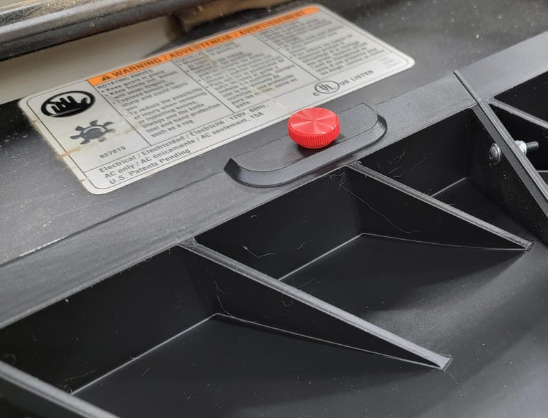 May include: Close-up of a black plastic surface with a red button. The surface has several compartments and a warning label with the text "A WARNING / ADVERTENCIA / AVERTISSEMENT" and "Electrical / Electricidad / AC seulement 120V 60Hz USA".