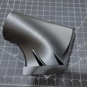 May include: A 3D printed black plastic part with a curved design. The part is sitting on a black cutting mat.