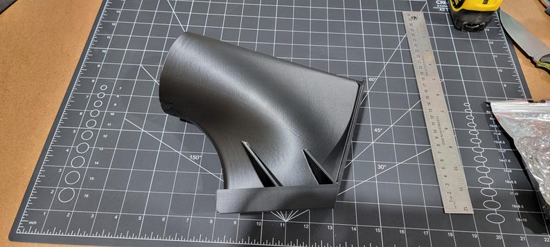 May include: A 3D printed black plastic part with a curved design. The part is sitting on a black cutting mat.