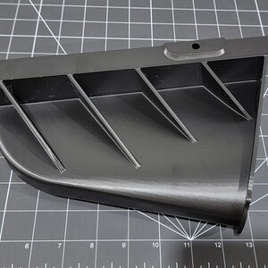 May include: A black plastic part with a series of angled fins. The part has a hole in the top and a rounded edge on the bottom.