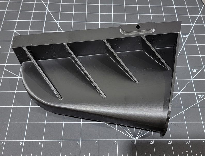 May include: A black plastic part with a series of angled fins. The part has a hole in the top and a rounded edge on the bottom.