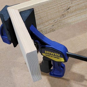 May include: A blue and black woodworking clamp with a yellow label that reads "IRWIN QUICK-GRIP" is used to hold two pieces of wood together at a 90-degree angle.