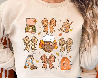 May include: Beige sweatshirt with a Thanksgiving-themed graphic. The design includes bows, a turkey chef, a lunchbox, a cutting board, an apron, and a milk carton with a cookie. The graphic is in shades of orange, brown, and beige.