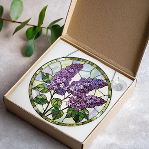 May include: A stained glass window featuring a lilac design, presented in a cardboard box. The round window showcases purple lilac blooms and green leaves, framed by a green border. A chain and suction cup are attached for hanging.