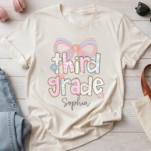 May include: A cream-colored t-shirt featuring a large graphic of a pink and blue bow above the text "third grade" in a playful, patterned font. Below the text, the name "Sophia" is printed in a black, cursive script font.