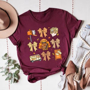 May include: Burgundy t-shirt featuring a collection of fall-themed illustrations. The design includes bows, mailboxes, a turkey dressed as a postal worker, and a mail truck. The shirt is adorned with autumn leaves, creating a festive and seasonal aesthetic.